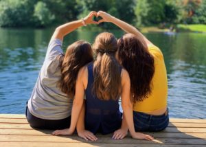 3 girls sitting by a body of water, lean in together to form a heart shape with their hands